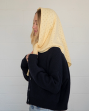 Person wearing a black jacket and light blue jeans with a beige headscarf against a white background