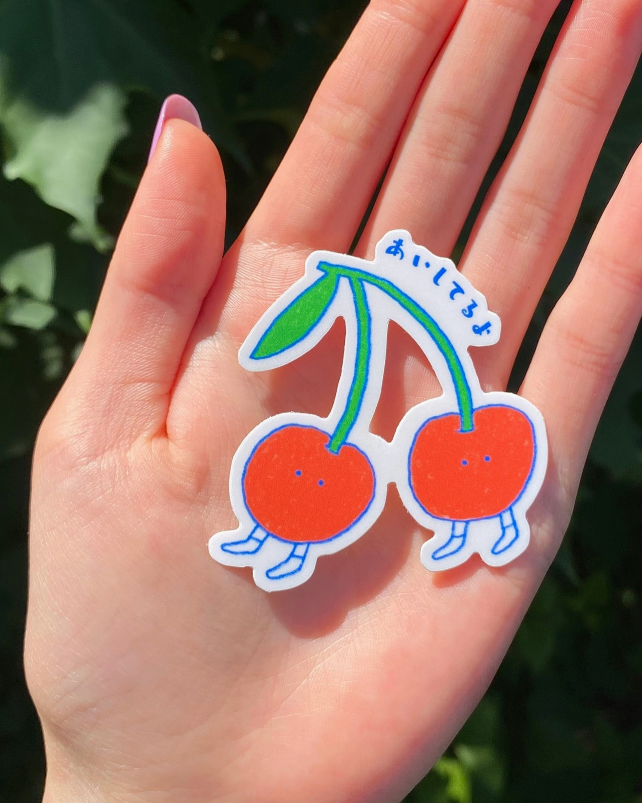 Hand holding two cartoon-style cherry stickers with a green leaf against a blurred natural background.