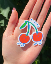 Hand holding two cartoon-style cherry stickers with a green leaf against a blurred natural background.
