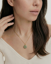 Woman wearing a gold necklace with a green pendant against a neutral background