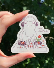 Hand holding a sticker of a cartoon character with headphones against a blurred green background
