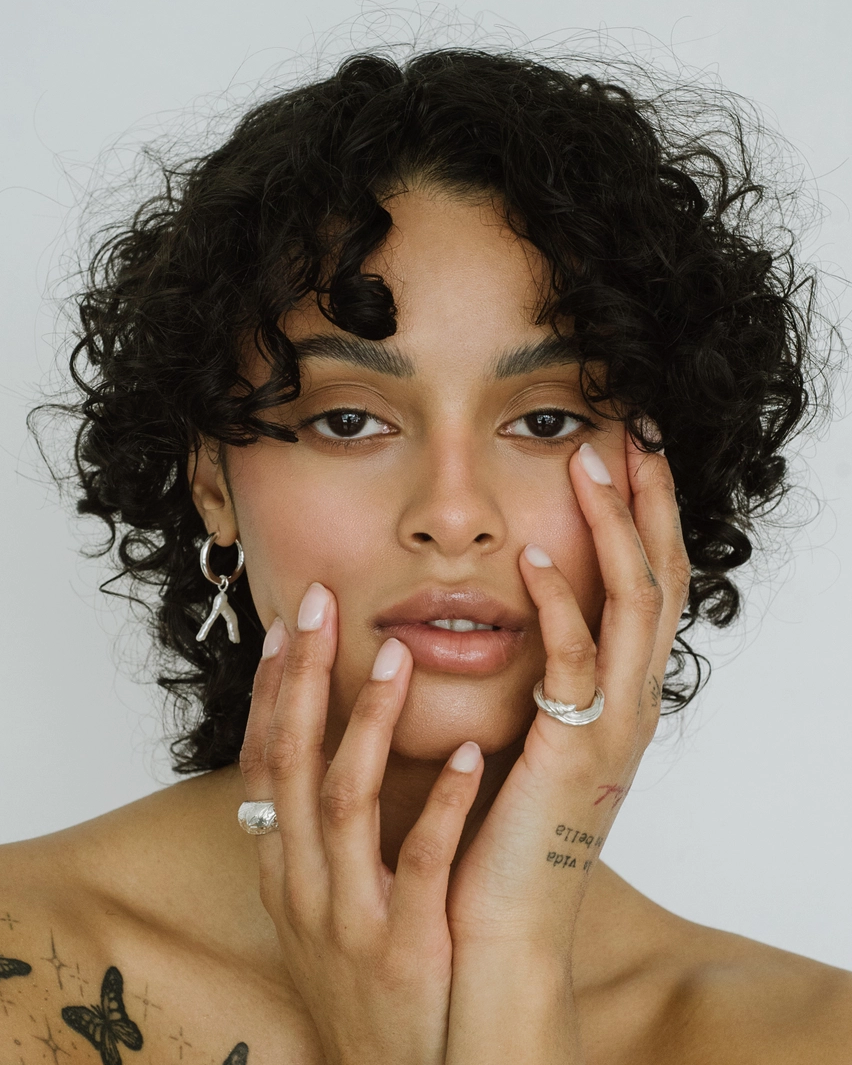 Woman with tattoos on her arms and chest, wearing earrings and a ring, against a plain background.