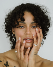 Woman with tattoos on her arms and chest, wearing earrings and a ring, against a plain background.