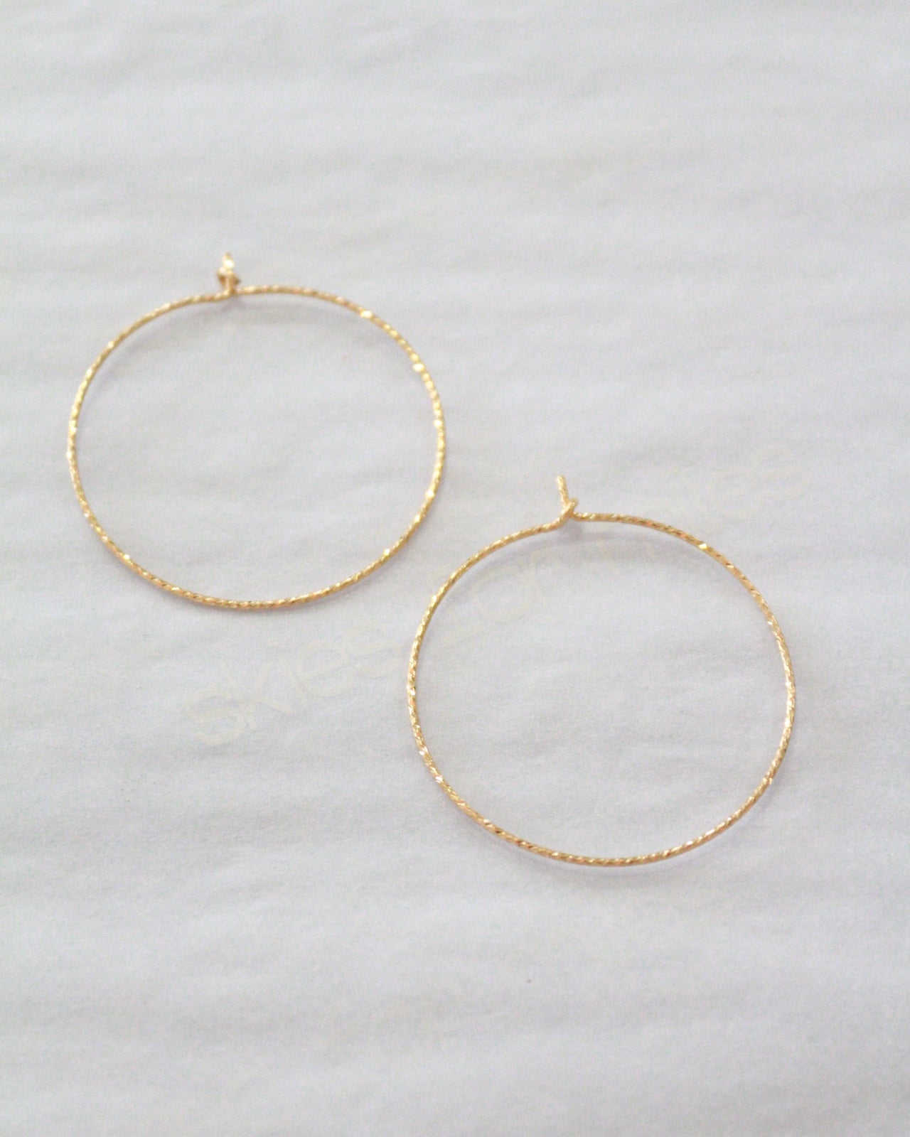 Two thin gold hoop earrings with a textured finish, displayed on a light gray fabric background.