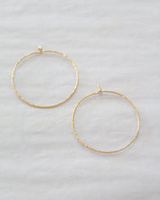 Two thin gold hoop earrings with a textured finish, displayed on a light gray fabric background.
