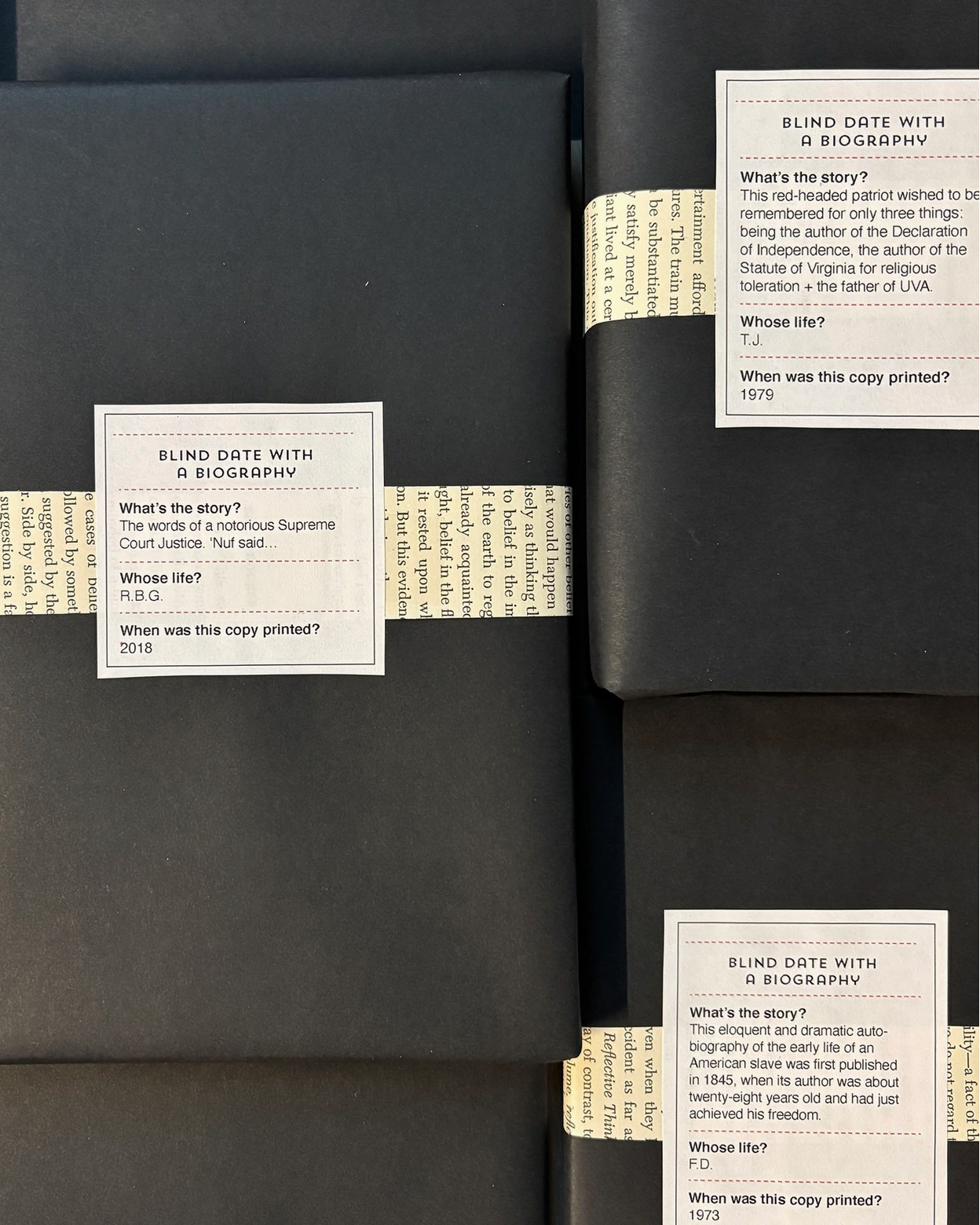 Several dark gray books wrapped in black paper with white labels are stacked closely together. The labels contain printed text and titles such as 'Blind Date with a Biography'.